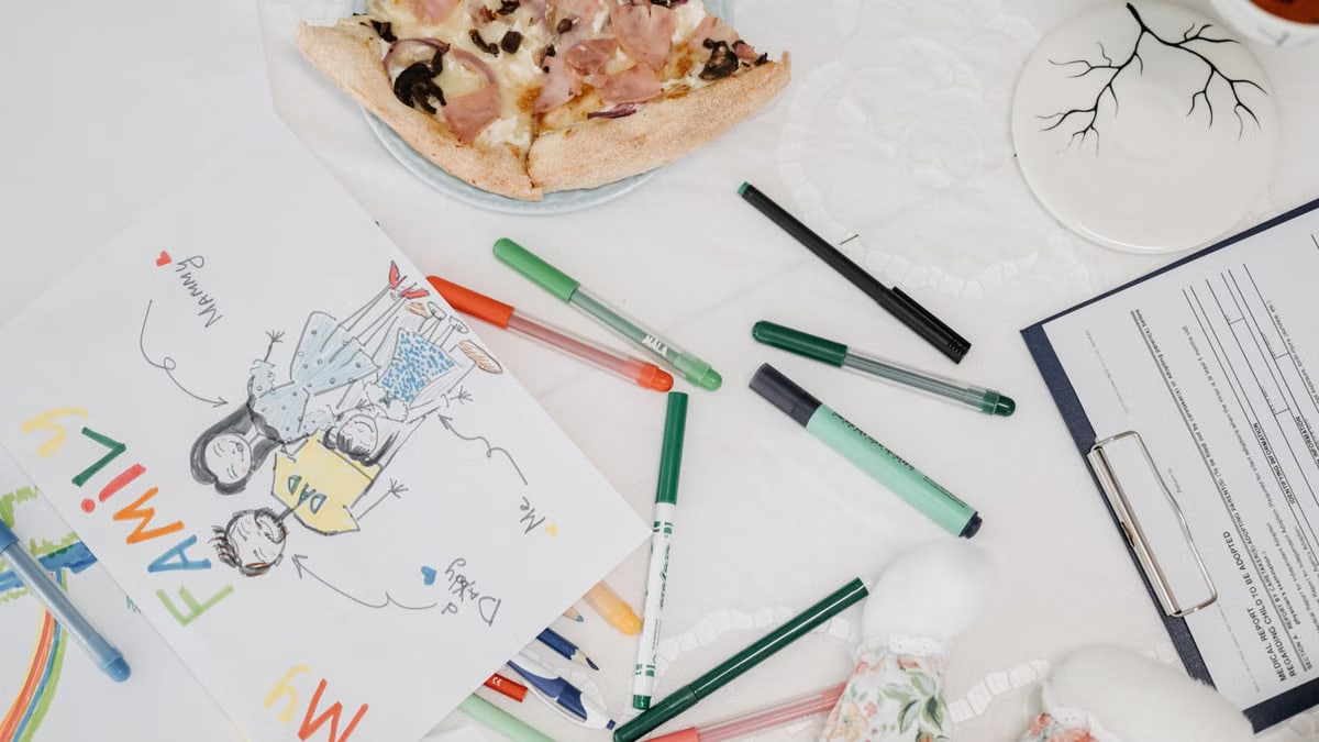 A colorful child's drawing of a family alongside pizza and pens on a table.